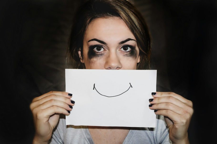 Frau hält ein Blatt mit einem lächelnden Smiley vor ihr Gesicht, wirkt dahinter aber ausgelaugt wegen dem dysfunktionalen System